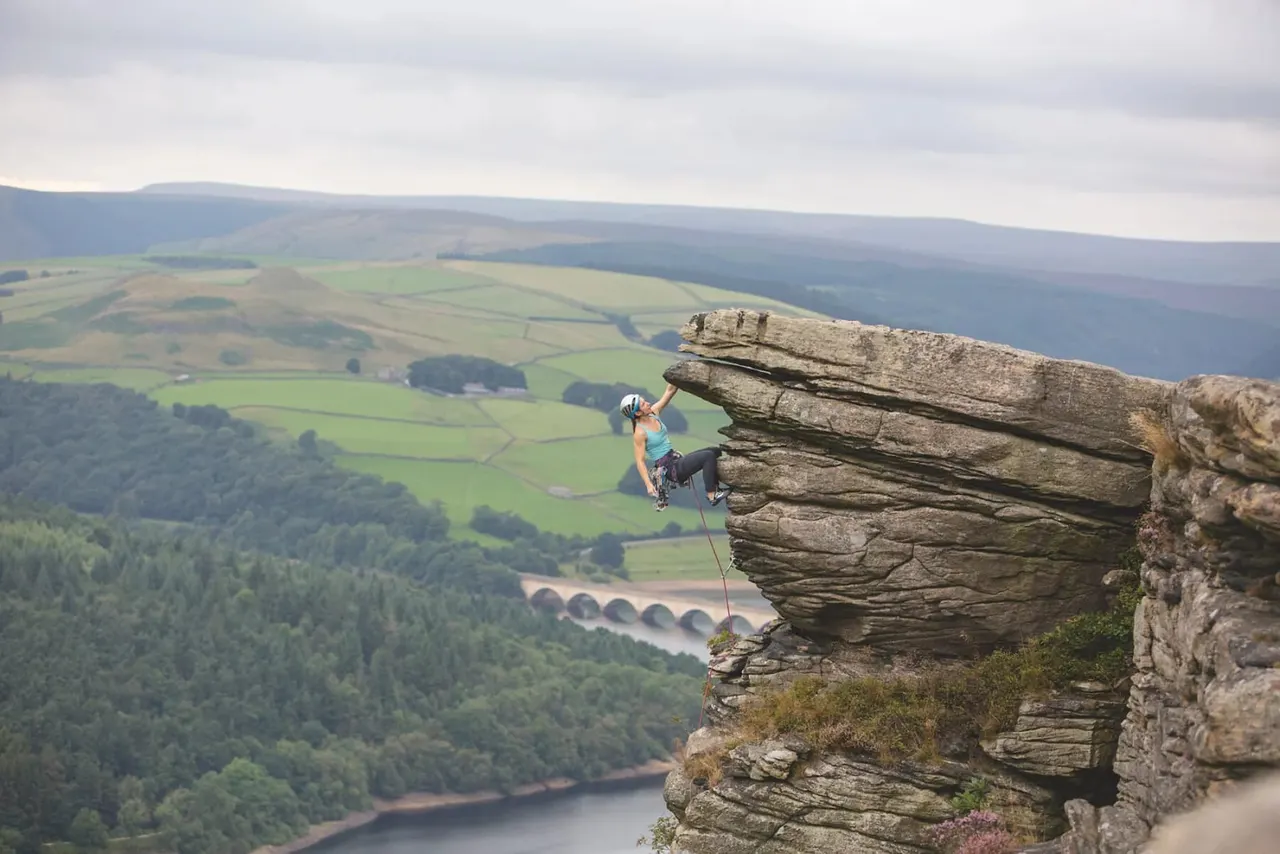Rock climber abseiling down a cliff face with expansive valley, forests and reservoirs visible below.