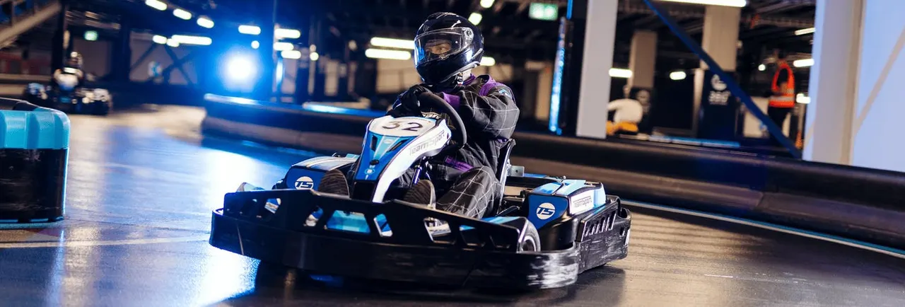 Person in helmet and racing suit driving a blue and white go-kart on an indoor track under blue lighting.