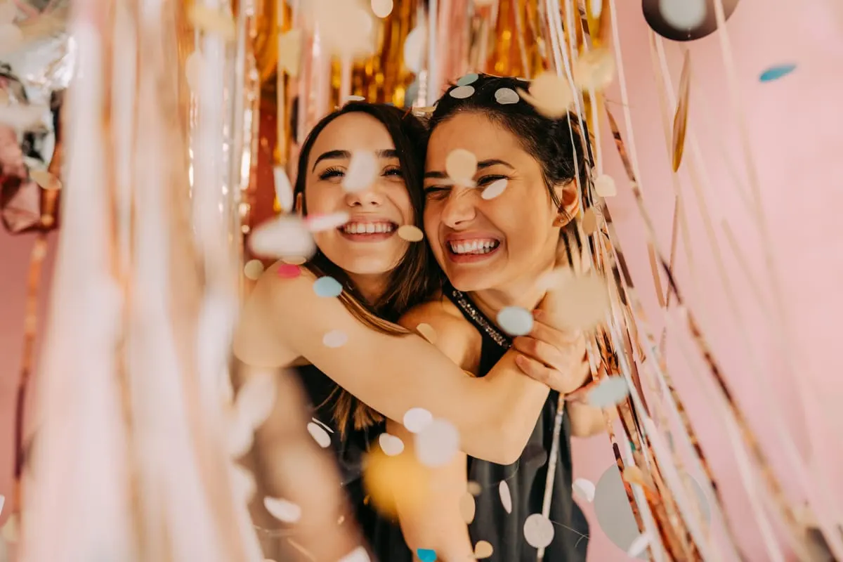 Two young women smiling and hugging surrounded by gold and white confetti streamers against a pink background.
