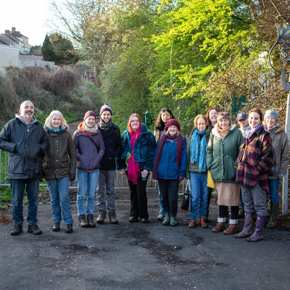A group of approximately fifteen adults stand together on a path, surrounded by lush green foliage and trees, wearing winter clothing.