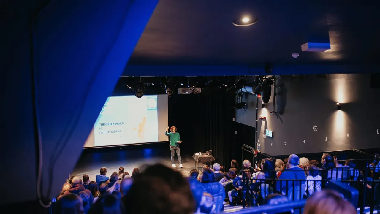 A speaker presents on stage to a seated audience in a theatre with a projected slide about Greek words visible on the screen.
