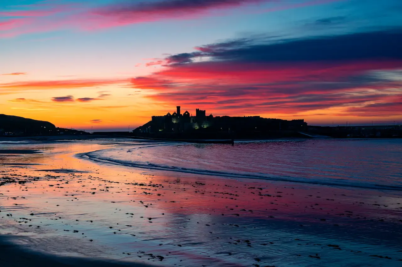 Sunset over a sandy beach with a silhouetted castle on an island across the water.
