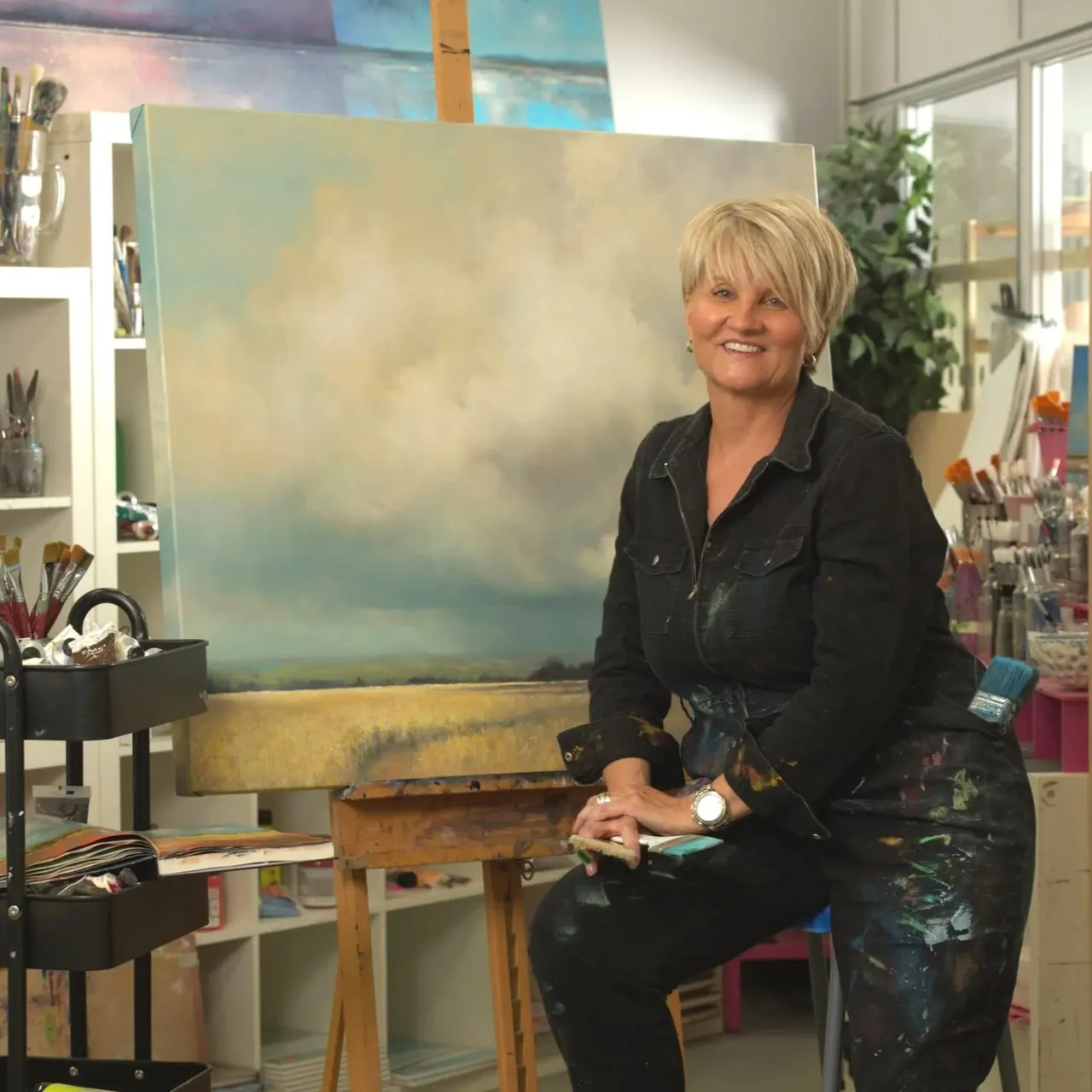 Woman sitting at easel in art studio with landscape painting and art supplies.