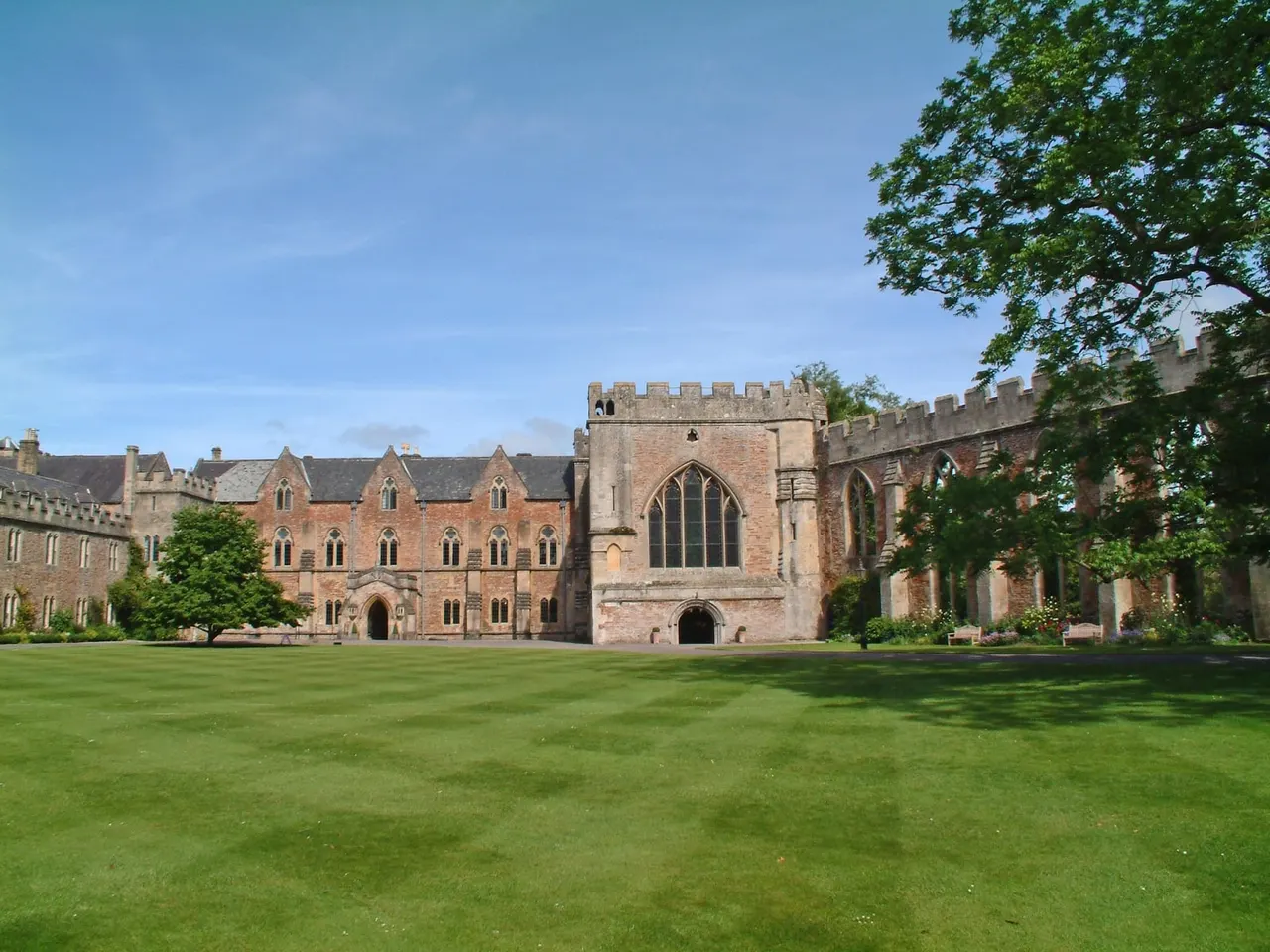 Medieval castle with crenellated towers and Gothic windows overlooks a wide green lawn on a sunny day.