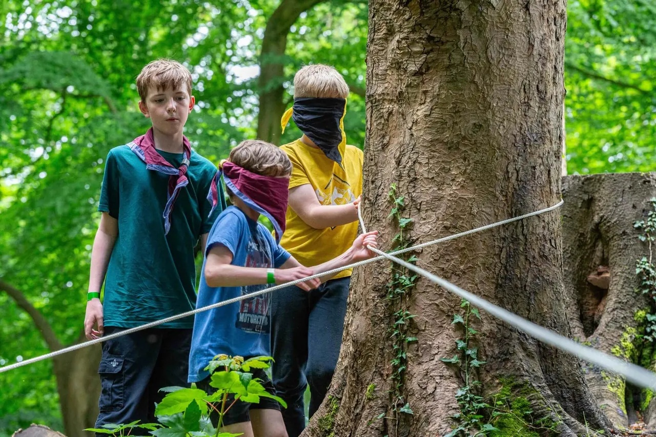 Three children in scout uniforms holding a rope course line next to a large tree in a woodland setting.