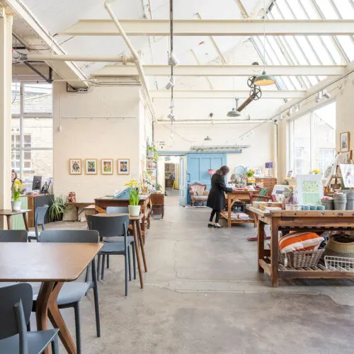 Bright studio or gallery space with high ceilings, wooden tables, chairs and potted plants, with one person walking through.