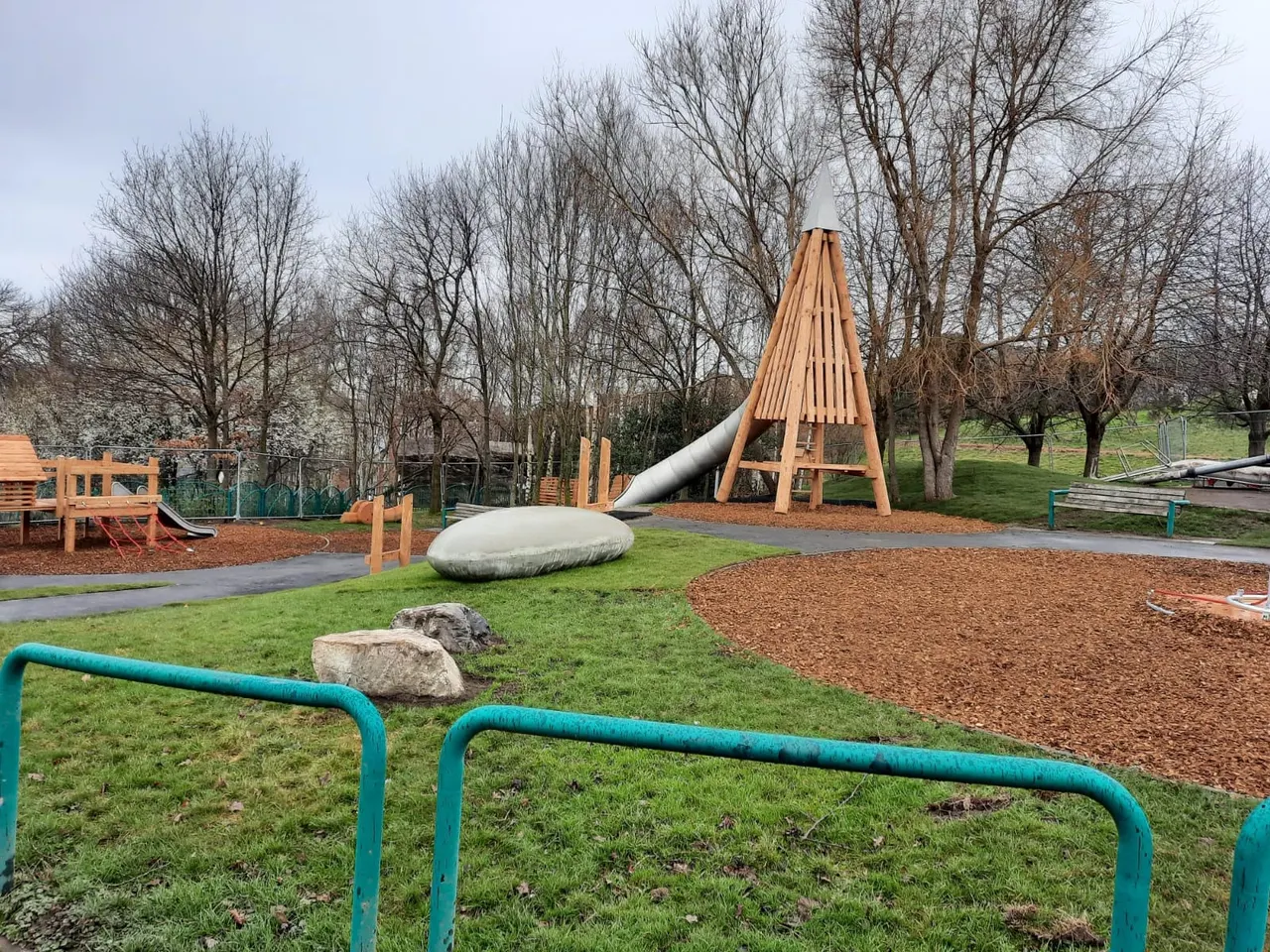 Modern playground with wooden climbing structures, slides, and equipment surrounded by bare trees and grass.