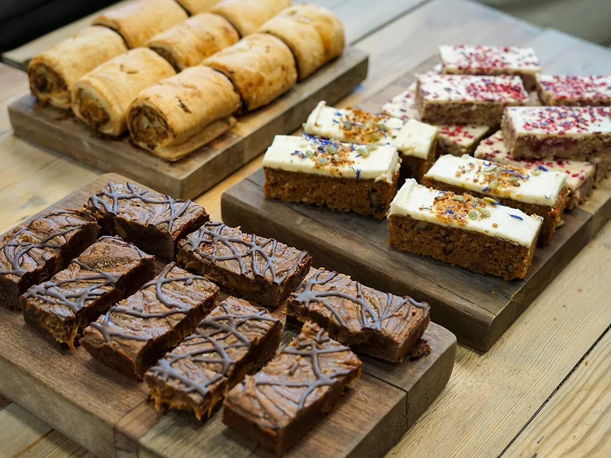 Selection of homemade cakes and pastries including chocolate brownies, frosted carrot cake slices and jam swiss rolls on wooden boards.