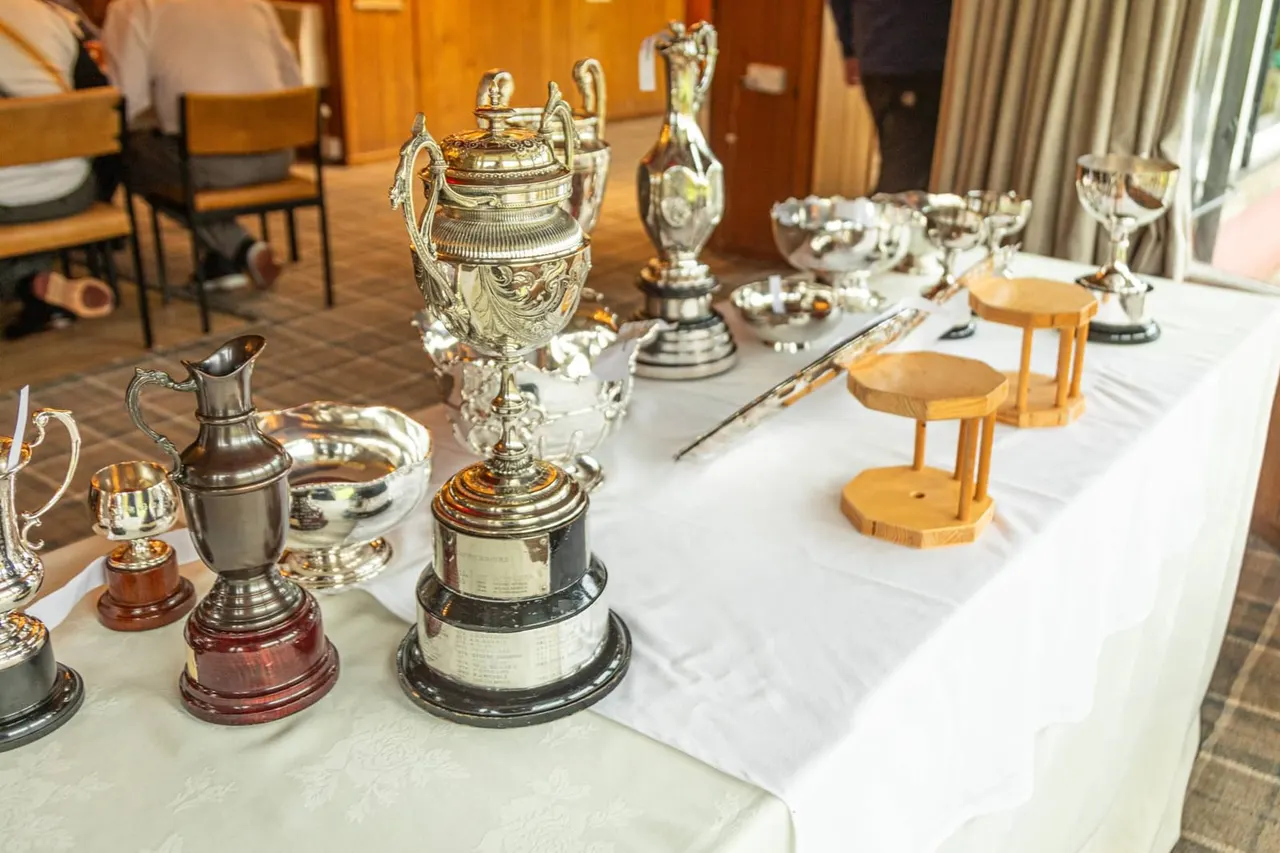 Collection of ornamental trophies and silverware displayed on a white-clothed table in a wood-panelled hall.