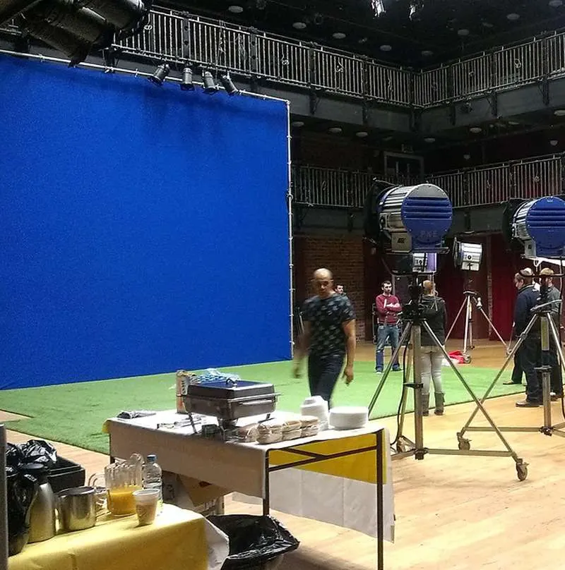 Film or television production studio with blue screen backdrop, green screen floor, professional lighting rigs, and crew members on set.