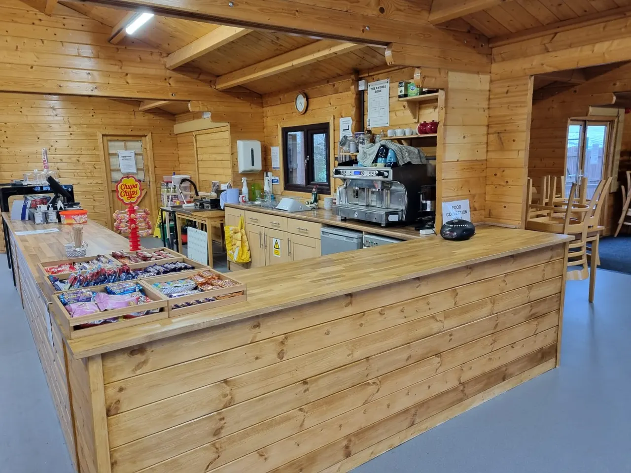 Interior of a wooden lodge with a service counter displaying confectionery and snacks, with professional catering equipment and espresso machine behind.
