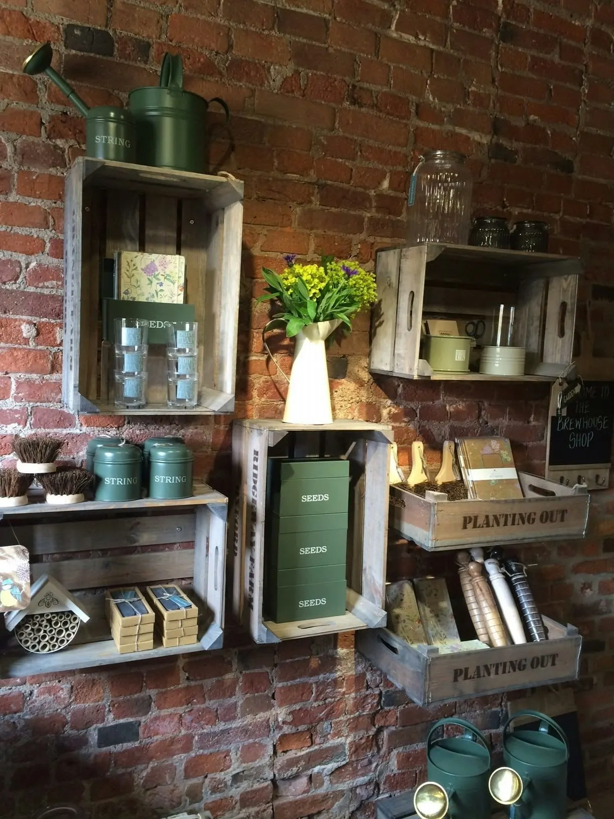 Garden shop display mounted on red brick wall with wooden shelves holding watering cans, seed packets, potted flowers and gardening tools.