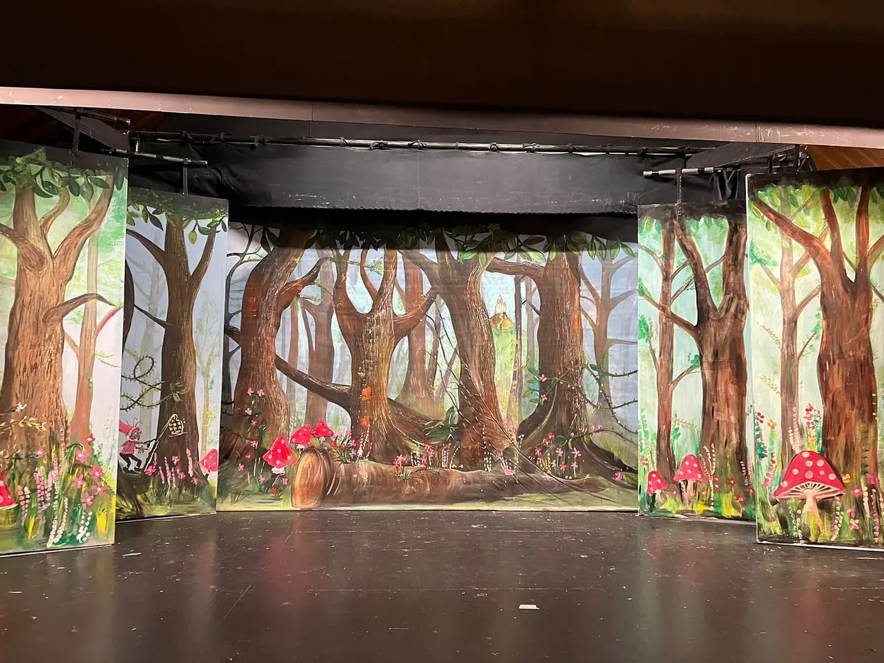 Theatre stage set depicting an enchanted woodland with painted backdrops of tall trees, red toadstools and flowers.
