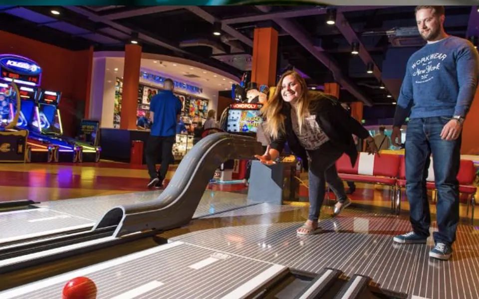 Woman bowling whilst man watches at an indoor leisure venue with arcade games and colourful lighting.