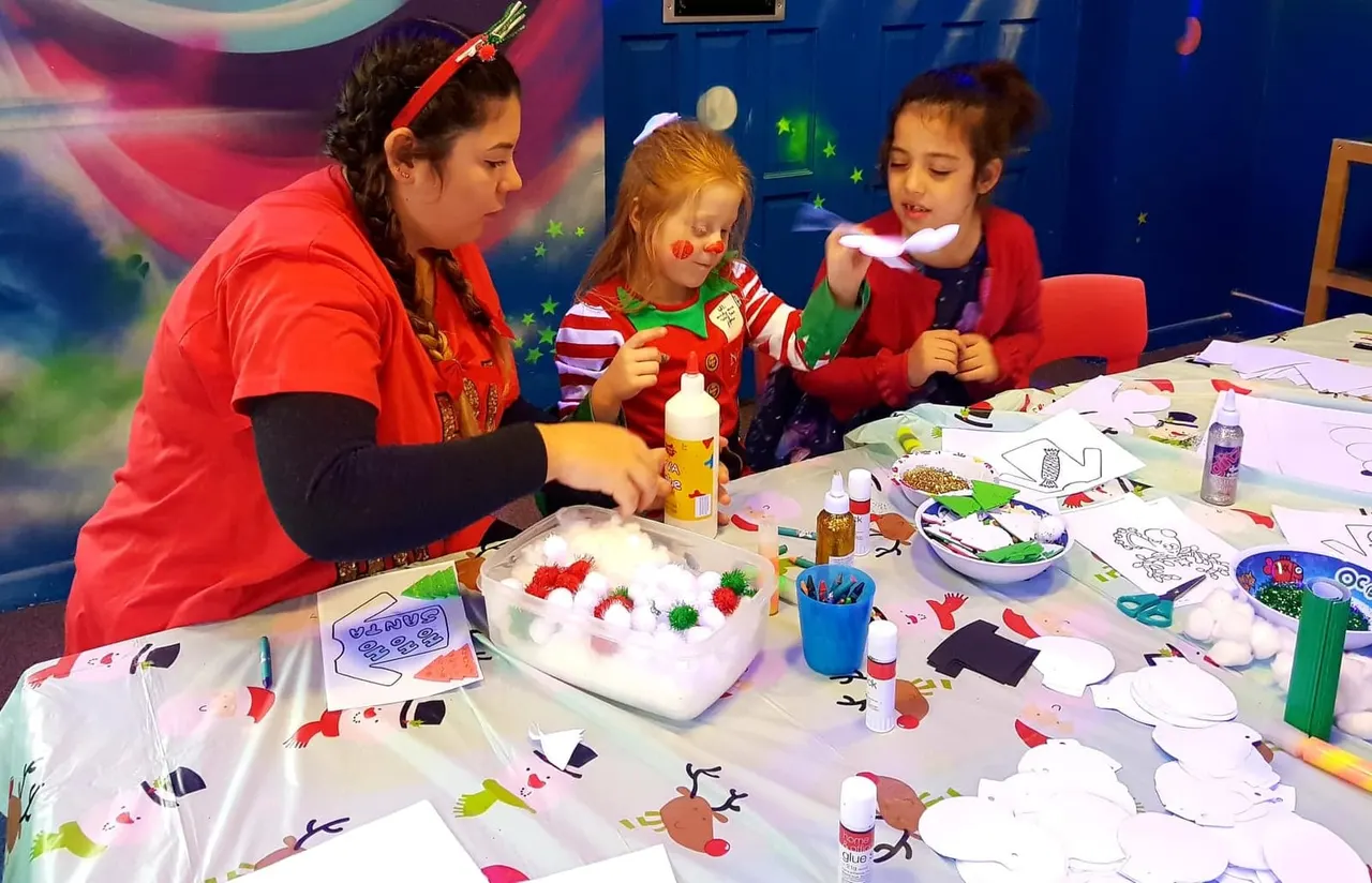 An adult staff member supervises two young children doing a festive craft activity at a table with coloured pom-poms, glue and decorations.