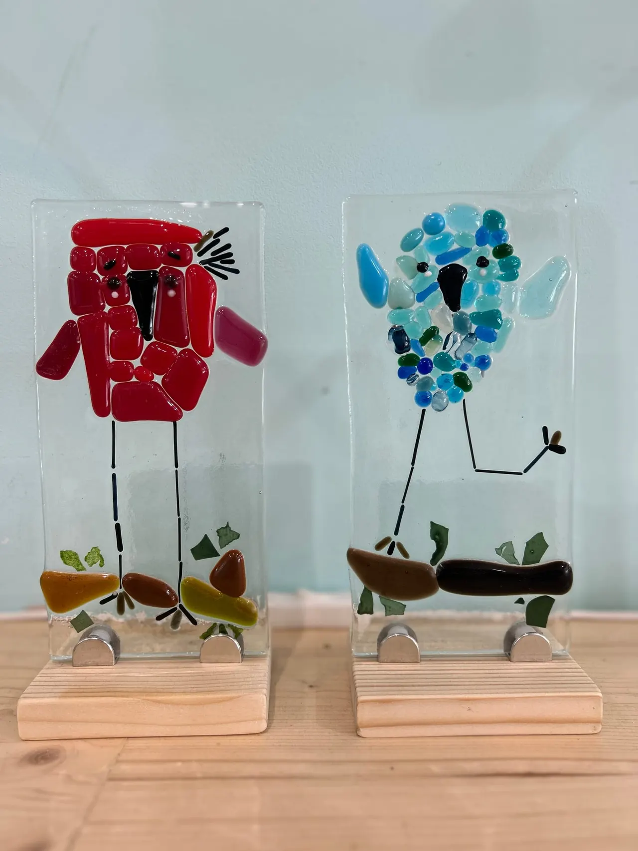 Two glass art pieces on wooden bases featuring stick-figure characters with coloured glass bodies and leaves, displayed on a wooden surface.