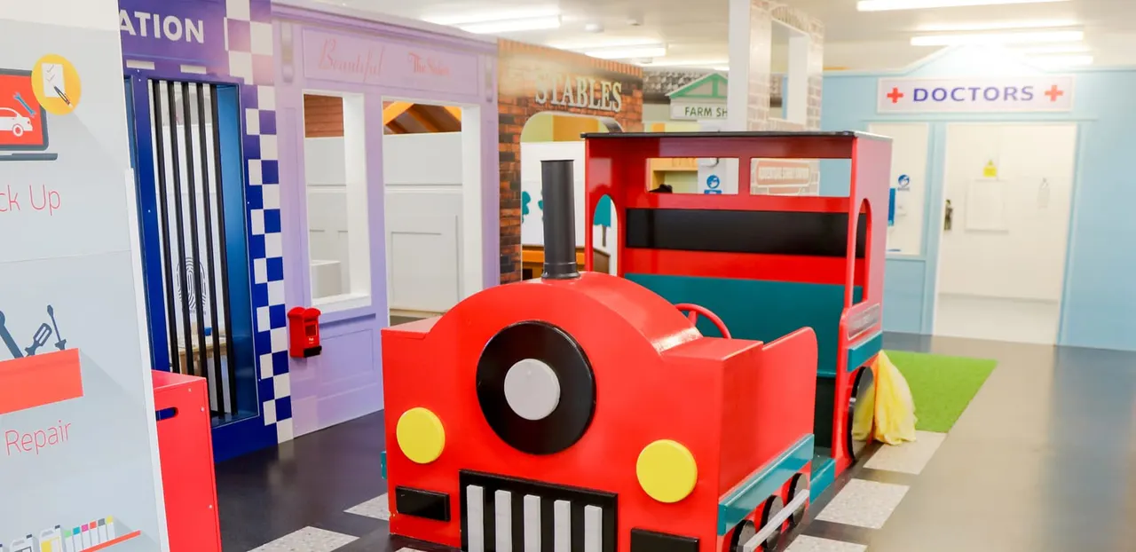 Indoor children's play area with colourful themed role-play zones including red vehicle and service station structures.