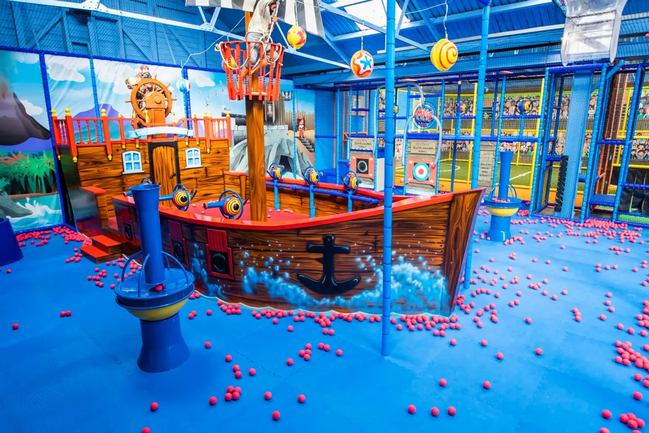 Indoor soft play area with pirate ship-themed structures, blue padded flooring covered in red balls, and climbing frames.