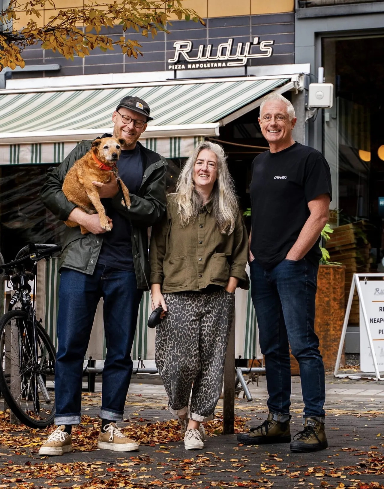 Three people and a dog stand smiling outside Rudy's Pizza Napoletana restaurant in autumn.
