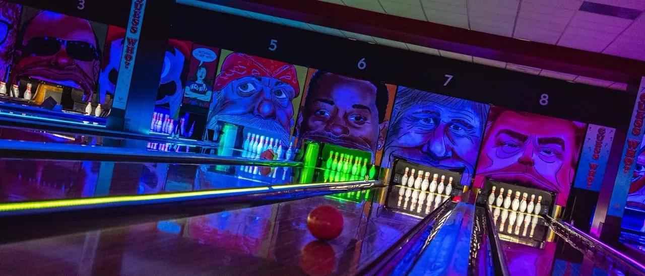 Bowling alley lane with neon lighting and colourful pop art portraits on the back wall behind the pins.