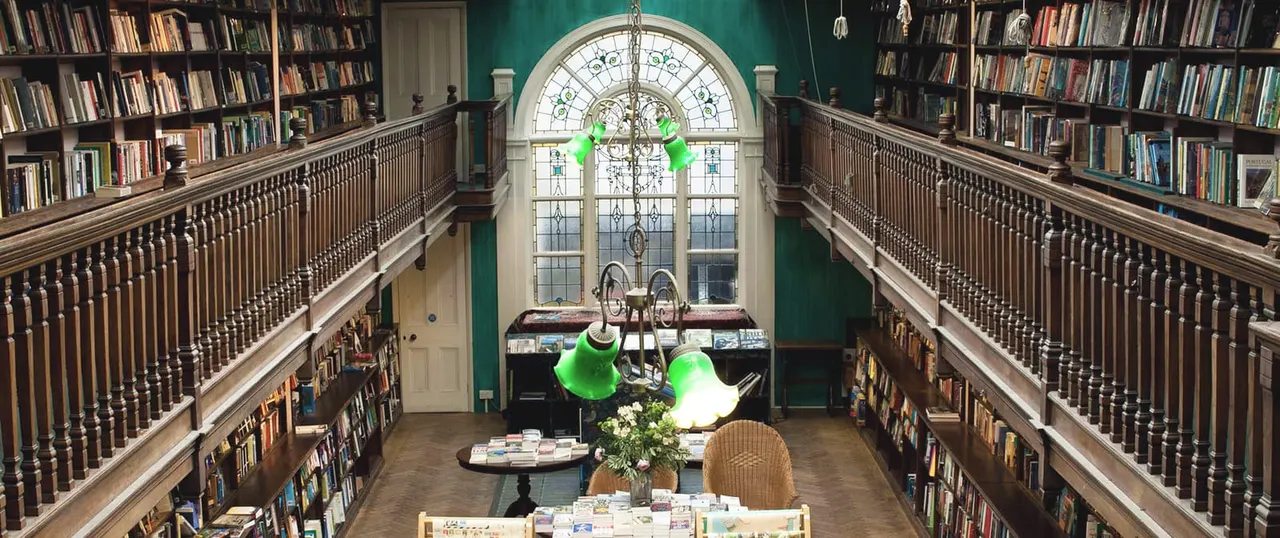 Interior view of a two-storey bookshop with green pendant lamps, ornate wooden railings, floor-to-ceiling bookshelves, and a large arched stained-glass window.