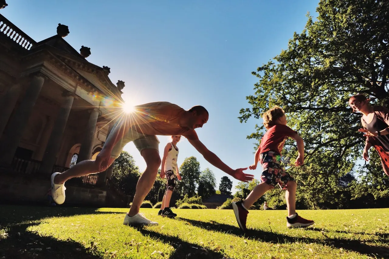 People playing tag on a sunny lawn next to a historic stone building surrounded by trees.