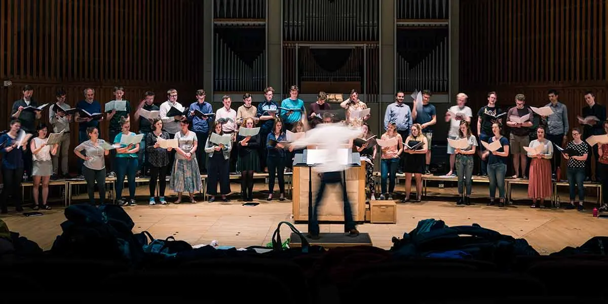 A large choir stands in rows on a concert hall stage, holding sheet music whilst a conductor leads them in performance.