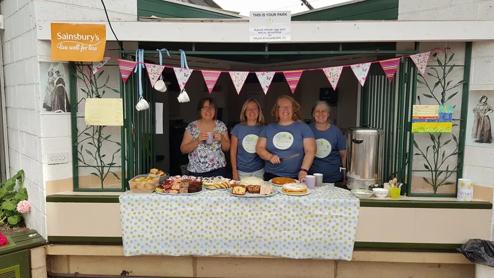 Four women staff a cake and bake sale stall at a park venue, with homemade goods displayed on a patterned tablecloth and bunting overhead.