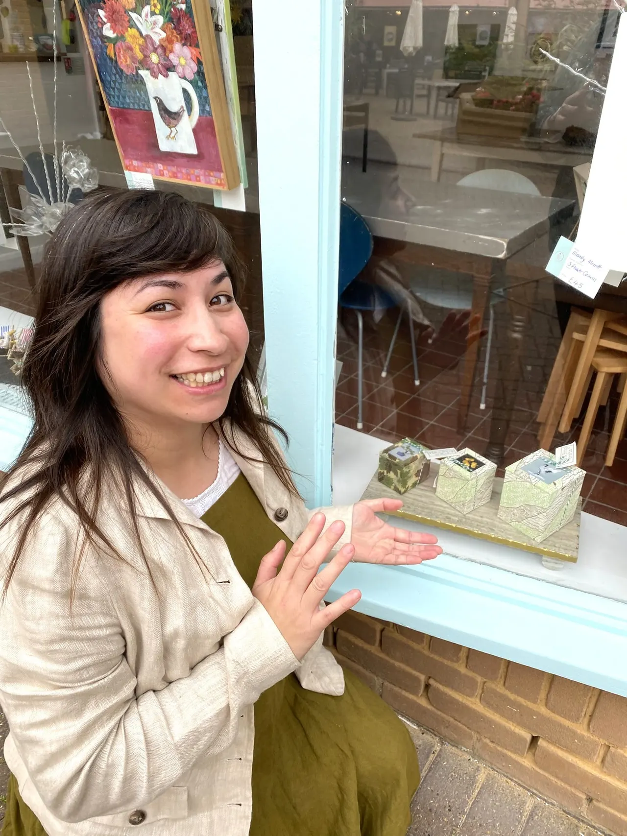 A smiling woman gestures towards a shop window display of decorative boxes and artwork inside a gallery.