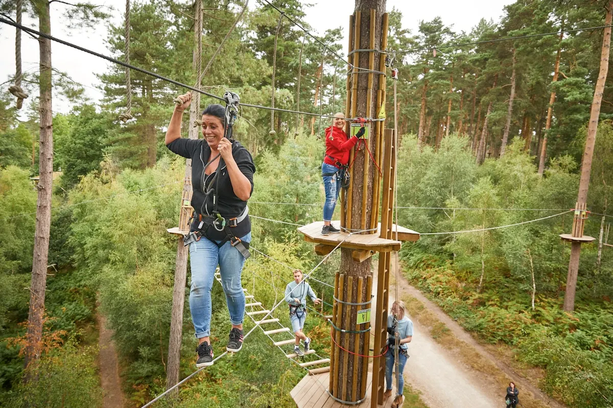 Woman on zip wire at forest adventure course with other participants on platforms among tall trees.