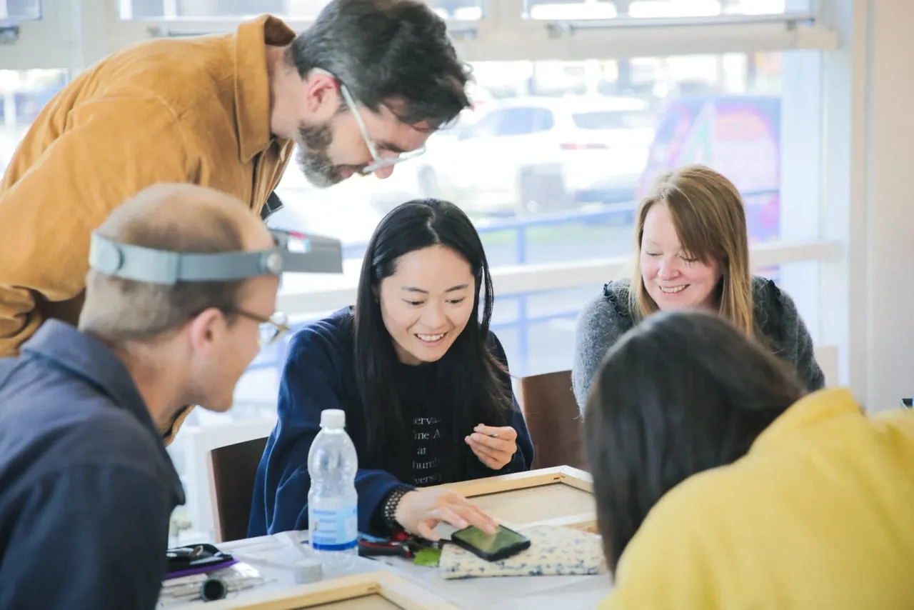 A diverse group of people gather around a table during an interactive workshop or educational session in a bright studio space.