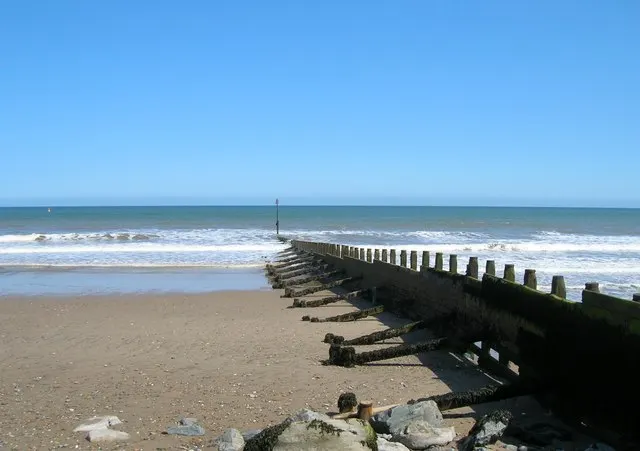 Wooden groynes extending into the sea along a sandy beach with waves and clear blue sky.