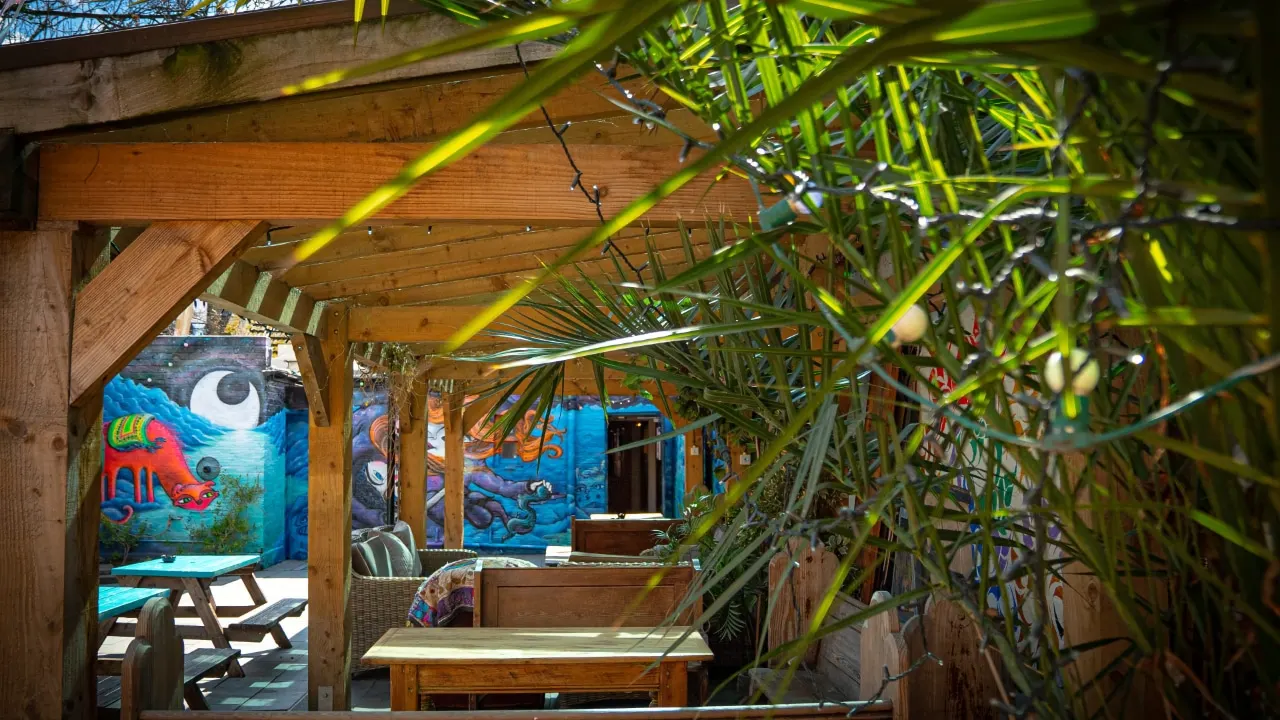Wooden pergola with hanging greenery framing a view of colourful murals and seating areas in an outdoor leisure space.