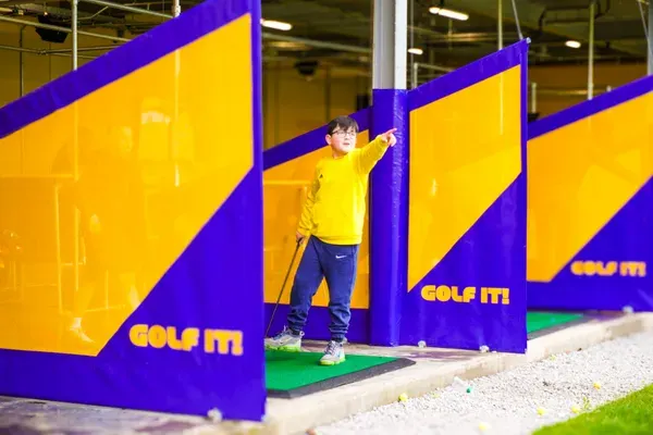 Child in yellow shirt putting on synthetic green at indoor mini golf course with blue and yellow obstacles.