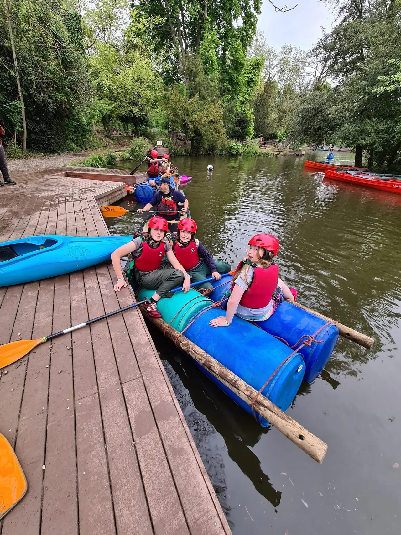 Children in red helmets and life jackets paddling colourful kayaks and canoes on a river lined with trees.