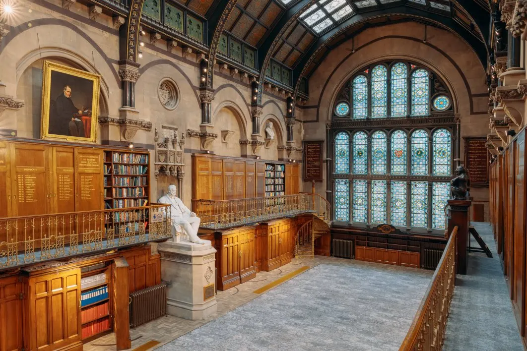 Interior of a grand historic library with ornate wooden panelling, arched ceilings, stained glass windows, bookshelves, a marble statue and portrait painting.