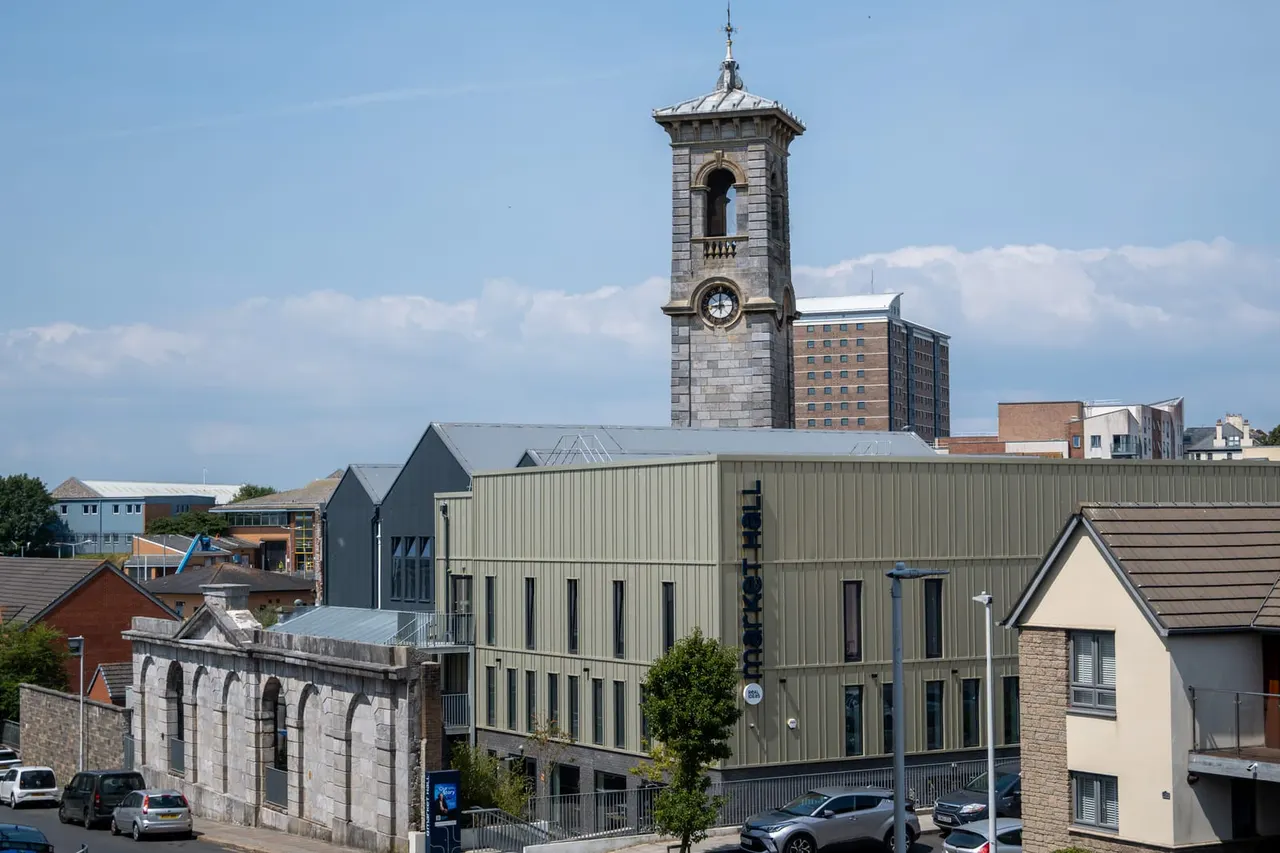 Townscape with historic stone tower and clock overlooking modern metal-clad building, residential properties and parked cars.