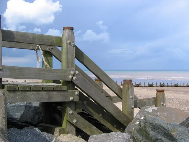 Wooden beach groyne with rocks and sandy beach stretching toward calm sea under blue sky.