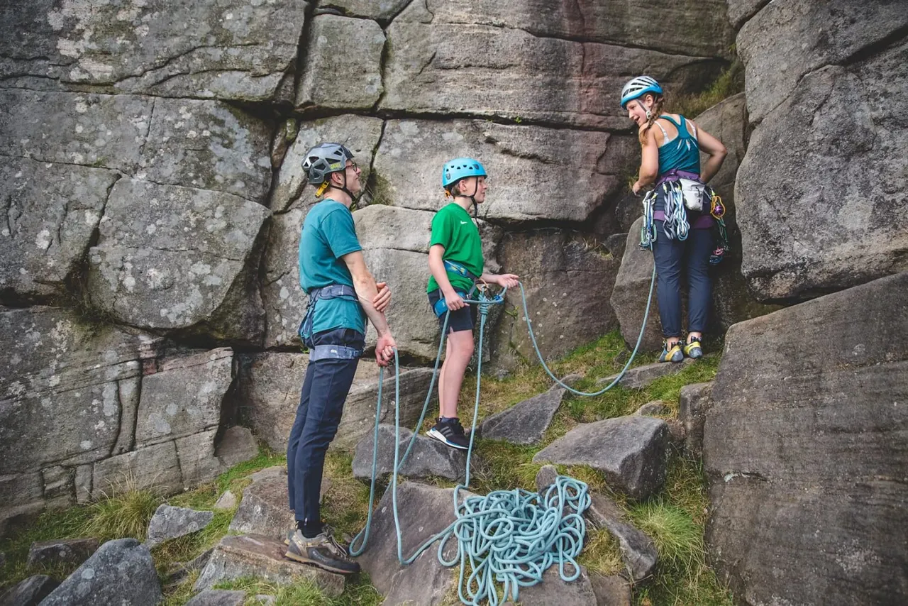 Three people wearing helmets and safety gear rock climbing against a large stone cliff face with ropes and equipment.