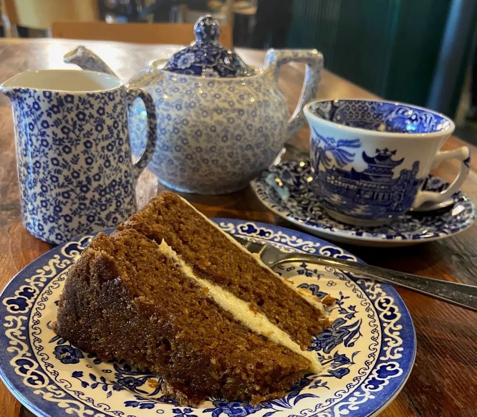 A slice of chocolate cake on a blue and white patterned plate with teapot, milk jug and teacup.