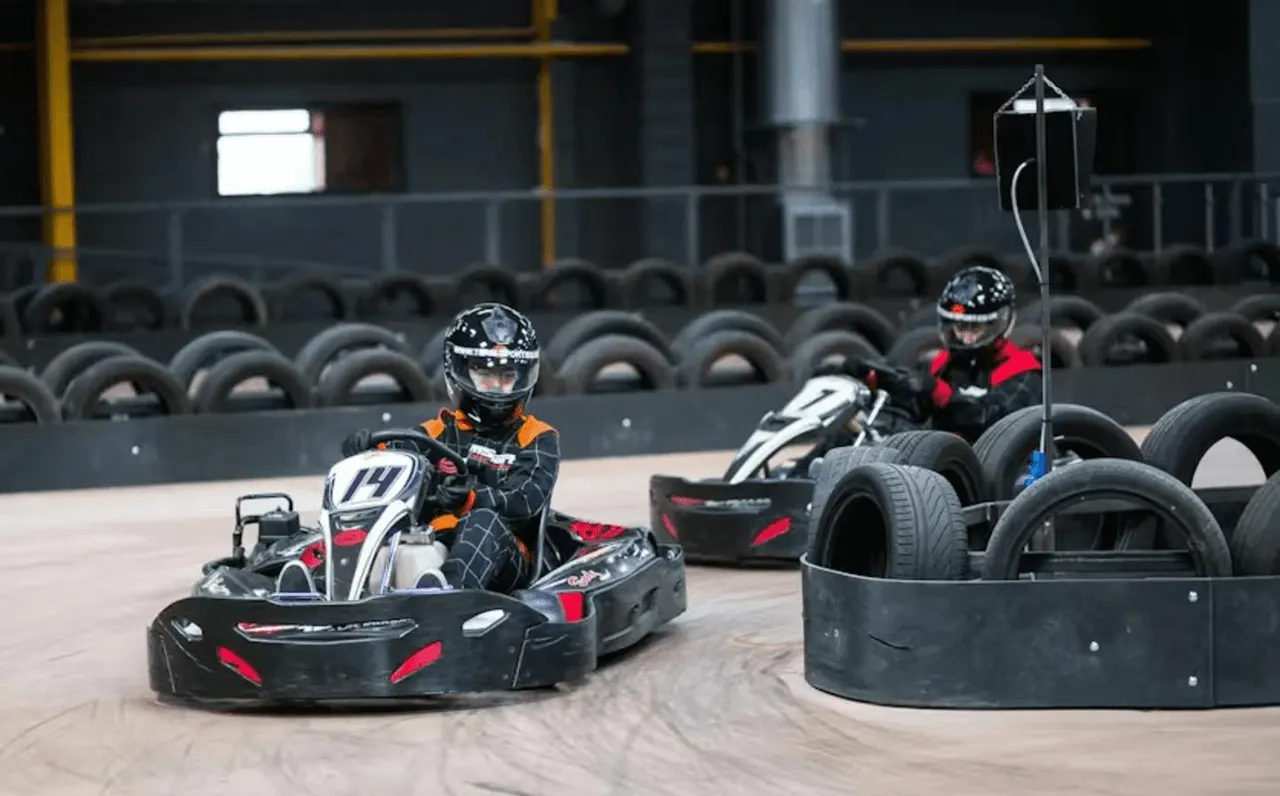 Two go-kart drivers in helmets racing on an indoor track with tyre barriers and a timing pole visible.
