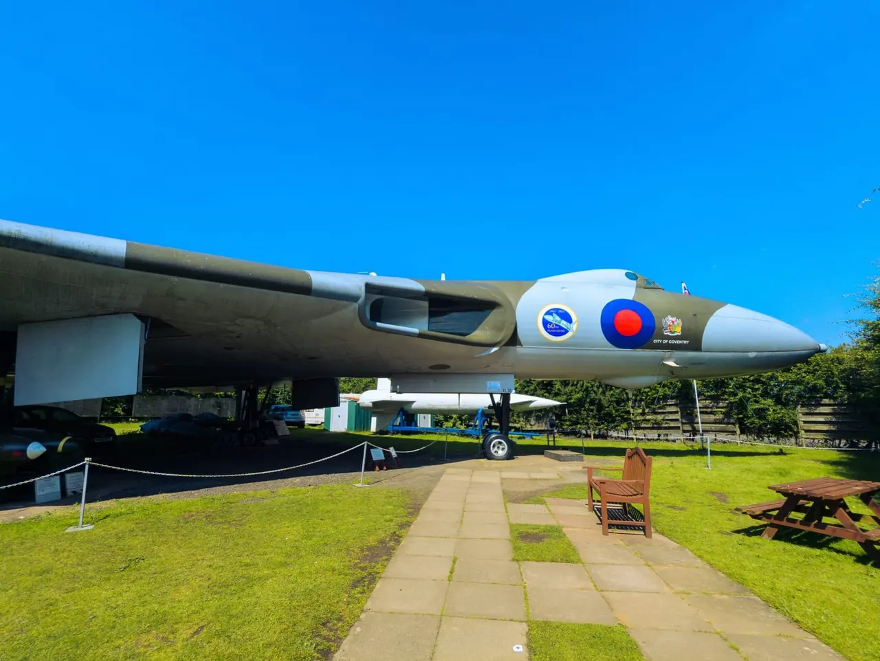 A retired military aircraft on display in a grassy outdoor setting with a paved pathway, picnic tables, and clear blue sky.