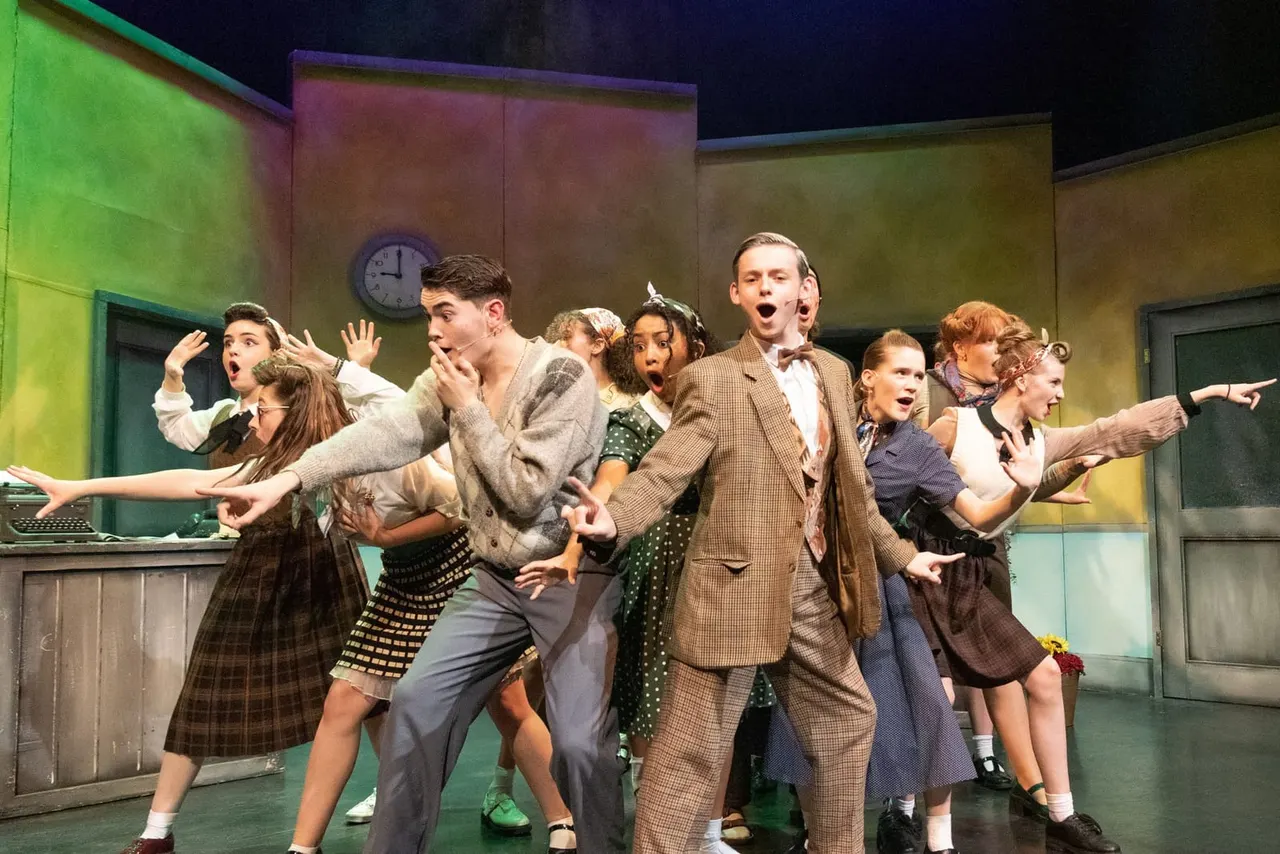 Cast of young performers singing and dancing together on a colourful lit stage set in 1950s style office interior.
