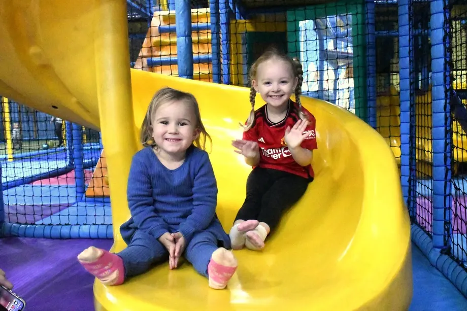 Two young children sliding down a bright yellow slide in an indoor soft play facility.