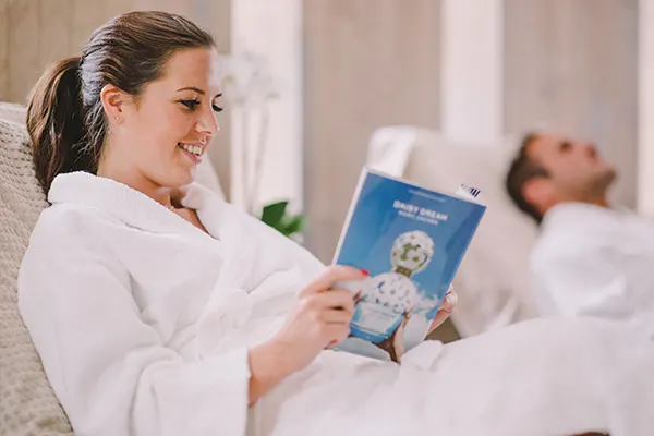 Woman in white robe reads blue magazine while relaxing on bed at spa venue.