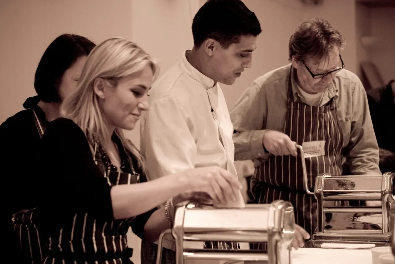 Four people in aprons and casual clothing gathered around a worktop, preparing food together in a cooking class.