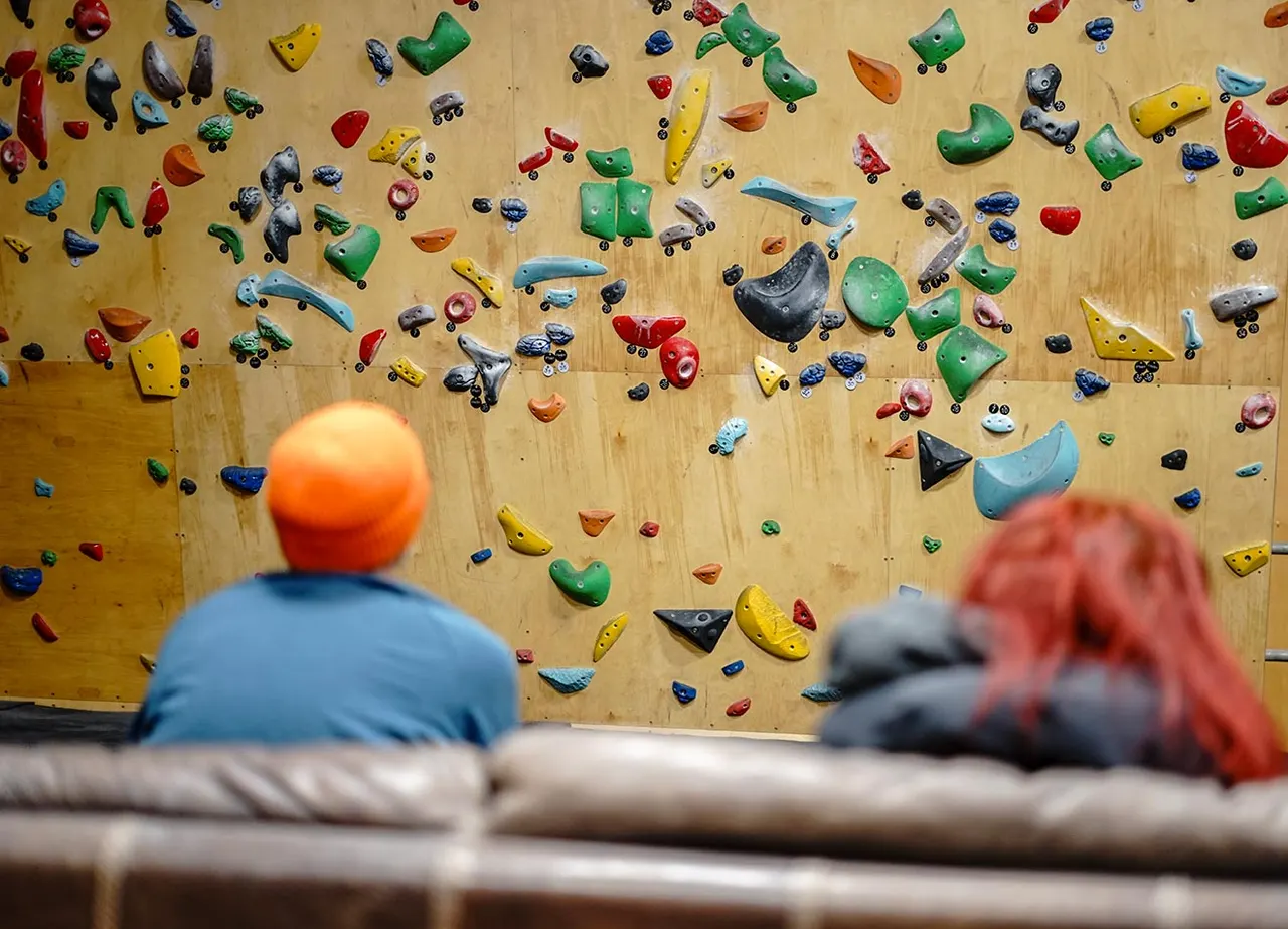 Two climbers in colourful helmets facing a wooden wall covered with multicoloured climbing holds.