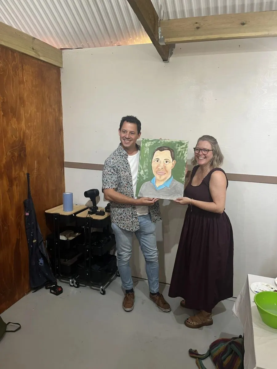 Two people hold up a portrait painting inside a rustic barn space with exposed beams.