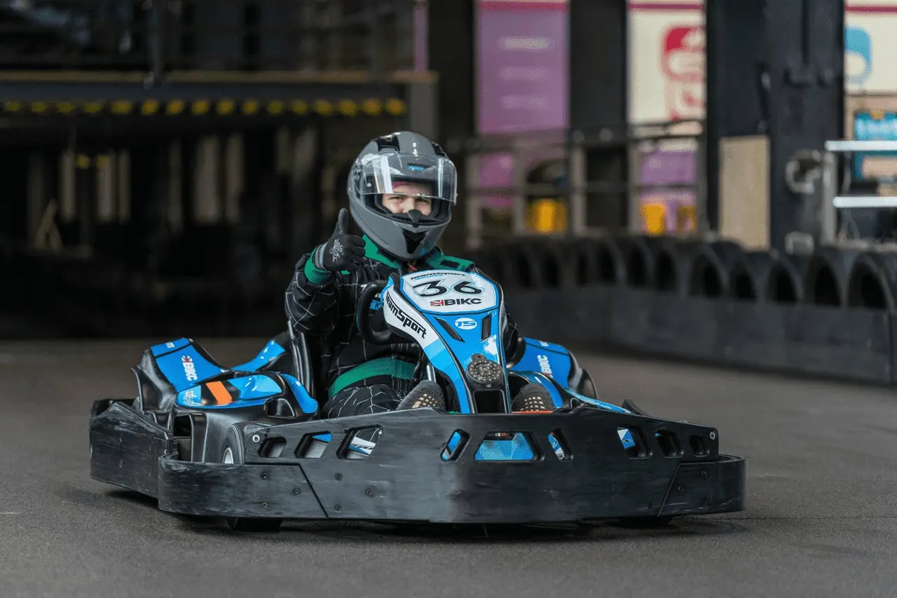Go-kart racer in helmet and protective gear driving a blue and black kart, giving a thumbs-up gesture on an indoor track.
