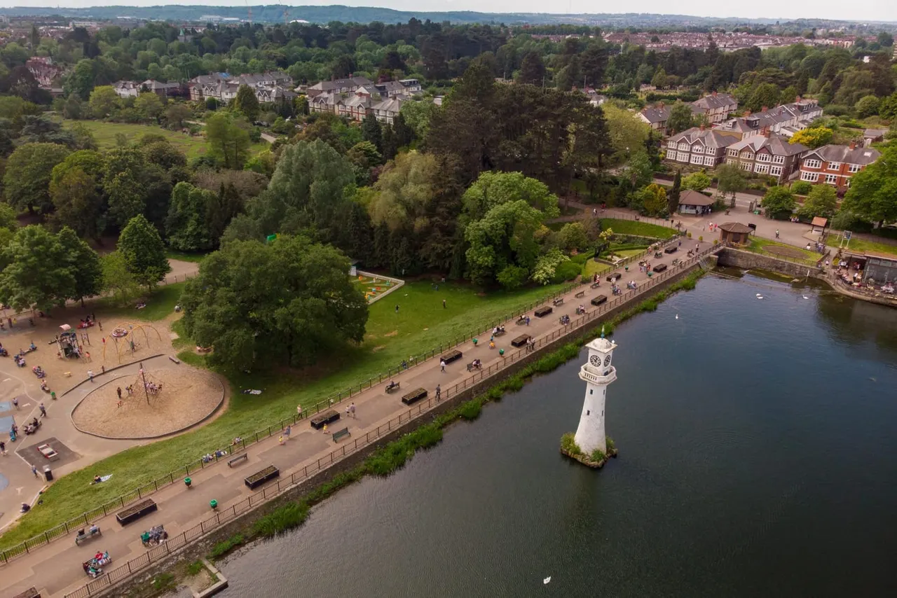 Aerial view of a riverside park with playground, waterside path, lighthouse, and surrounding suburbs with trees and hills beyond.